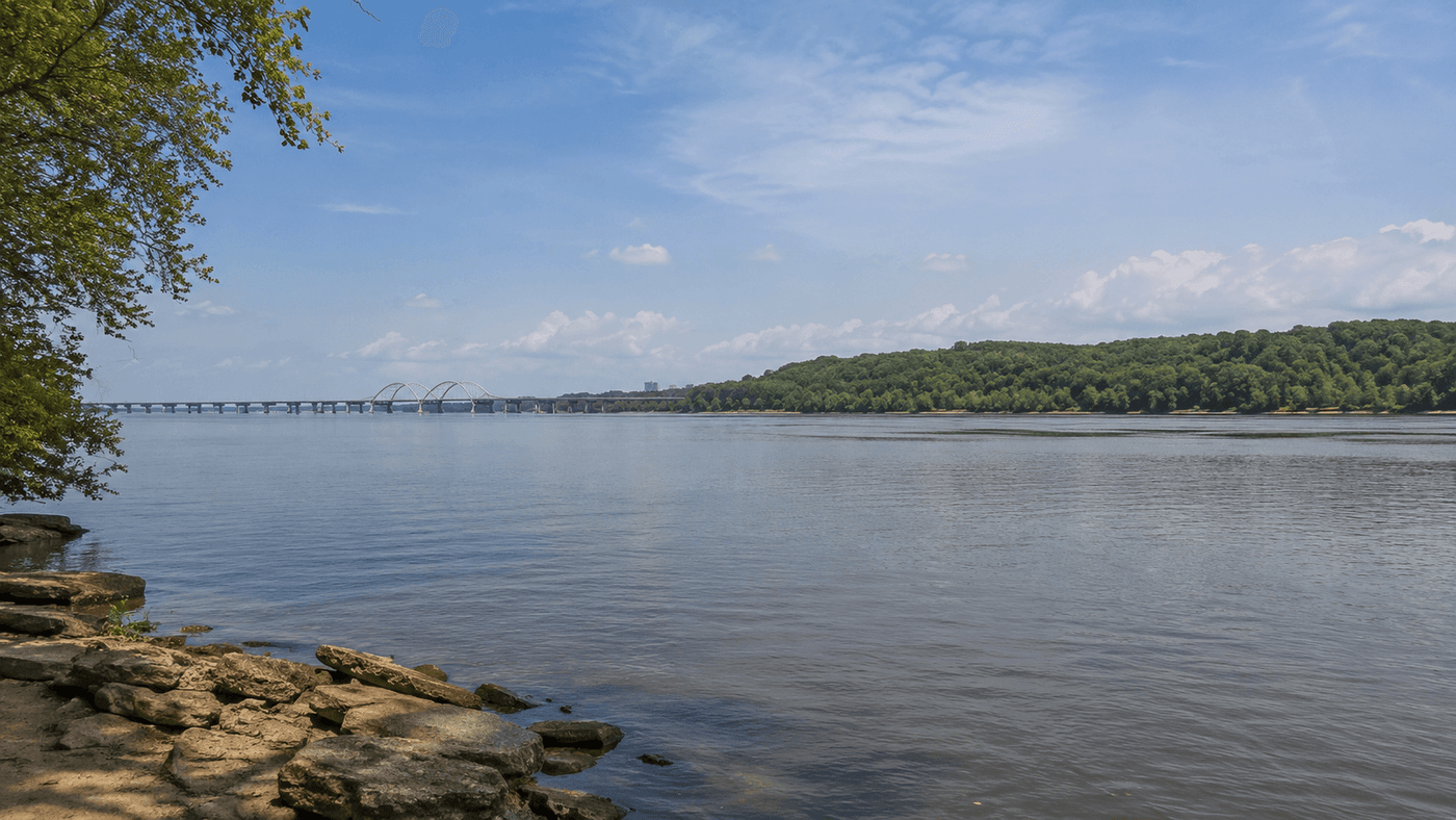 The Potomac River near Fort Washington, Maryland, with the wooded Maryland shoreline and the Woodrow Wilson Bridge visible in the distance toward Alexandria, Virginia.