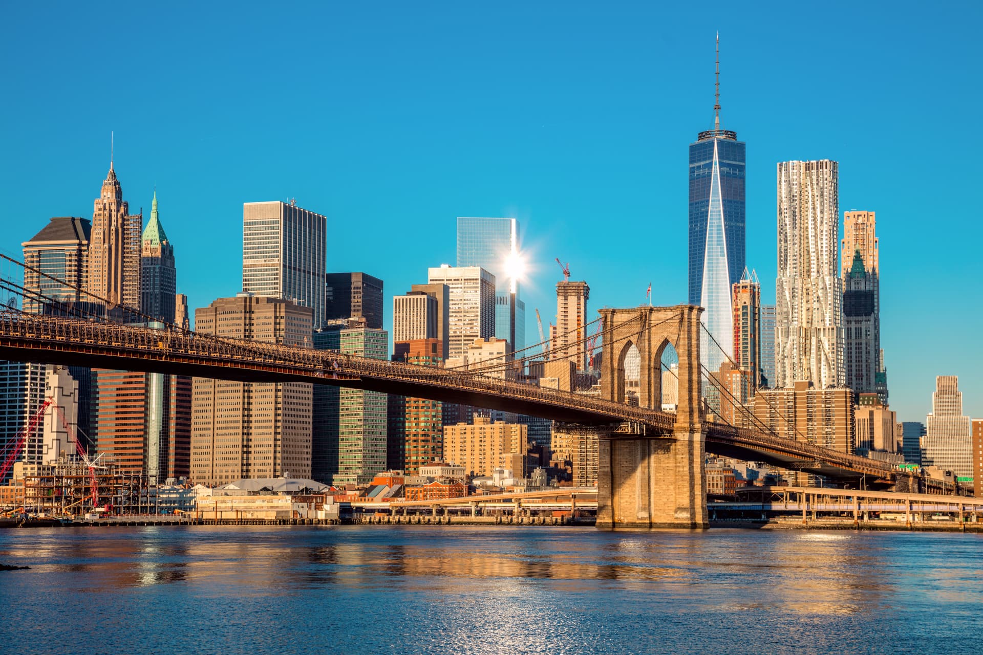 The Lower Manhattan skyline viewed from across the water, with the cluster of historic and modern towers in the Financial District under a clear sky.