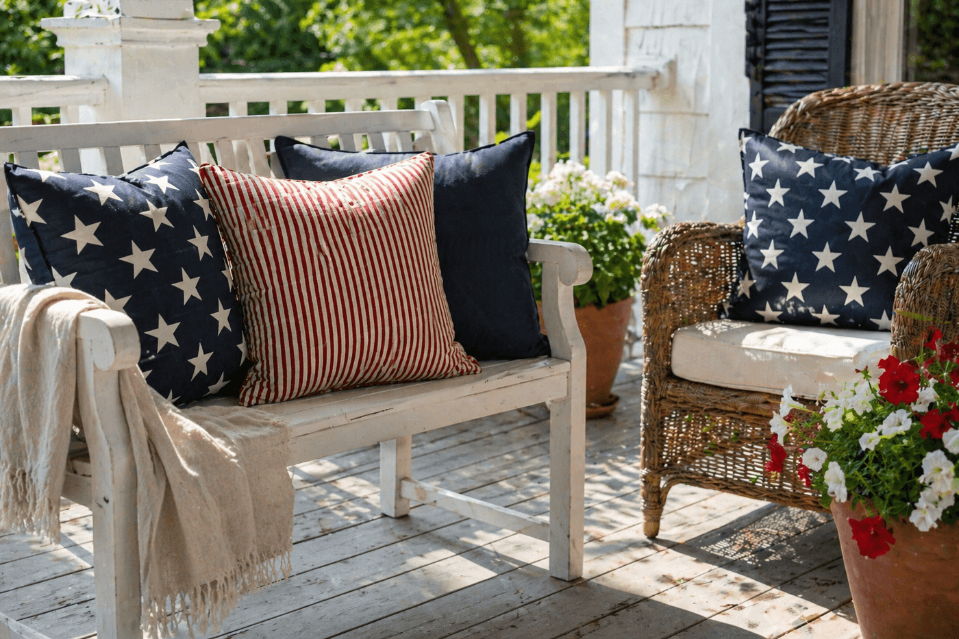Four patriotic throw pillow covers in coordinating star, stripe, and solid navy prints arranged on a white porch bench and wicker chair, styled with a small potted plant and neutral linen throw