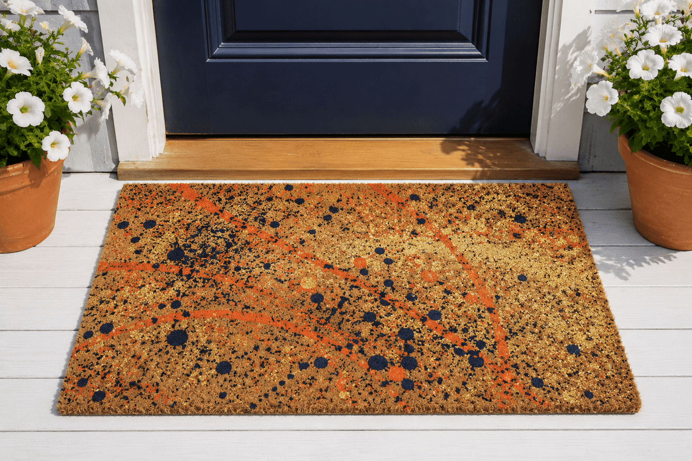 Finished splatter-painted coir doormat with layered fine speckle and bold arc paint splatters in navy, coral, and gold on natural coir fiber, displayed at a front door entry with potted plants flanking it