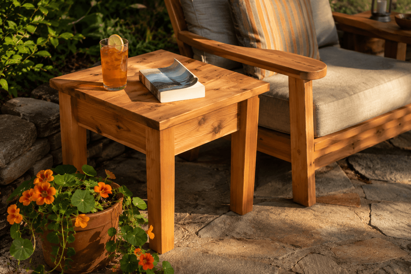 A handbuilt cedar outdoor side table beside a patio lounge chair holding a sweating glass of iced tea and a paperback book, natural wood grain visible in warm afternoon sun