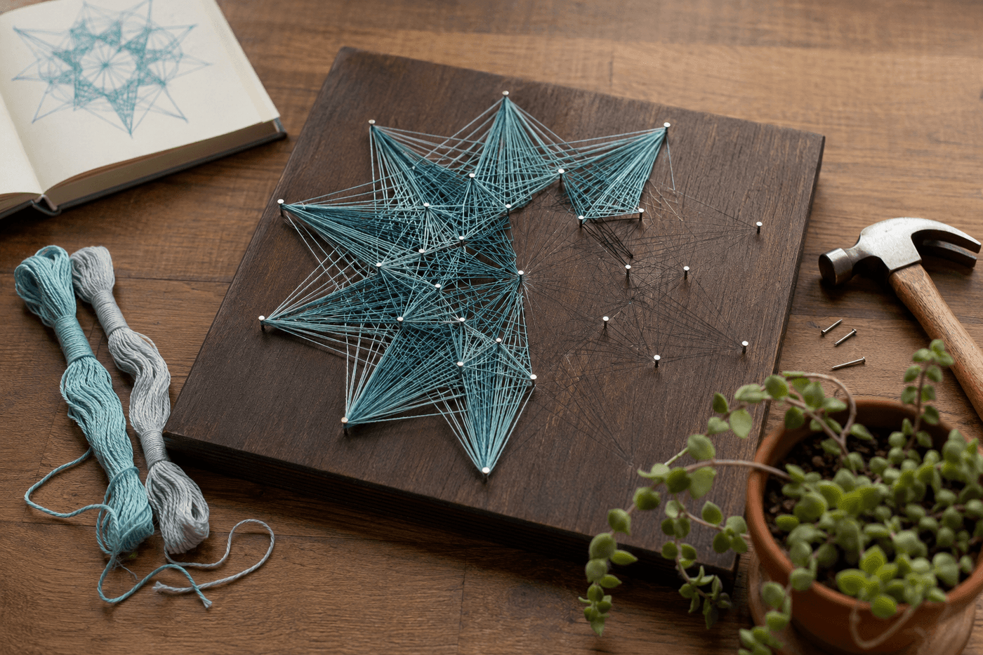 A finished string art piece on a dark stained wooden board showing a geometric star design in soft blue and teal embroidery floss with small silver nail heads visible, beside a small hammer and remaining floss on a wooden desk