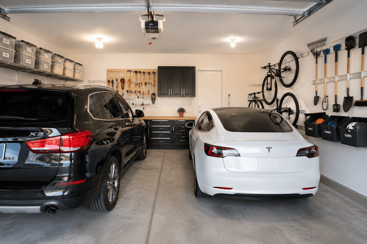 A fully organized two-car garage with wall-mounted track system holding bikes, long-handled tools, and sports bins, swept concrete floor with two cars parked inside and clear dedicated zones visible