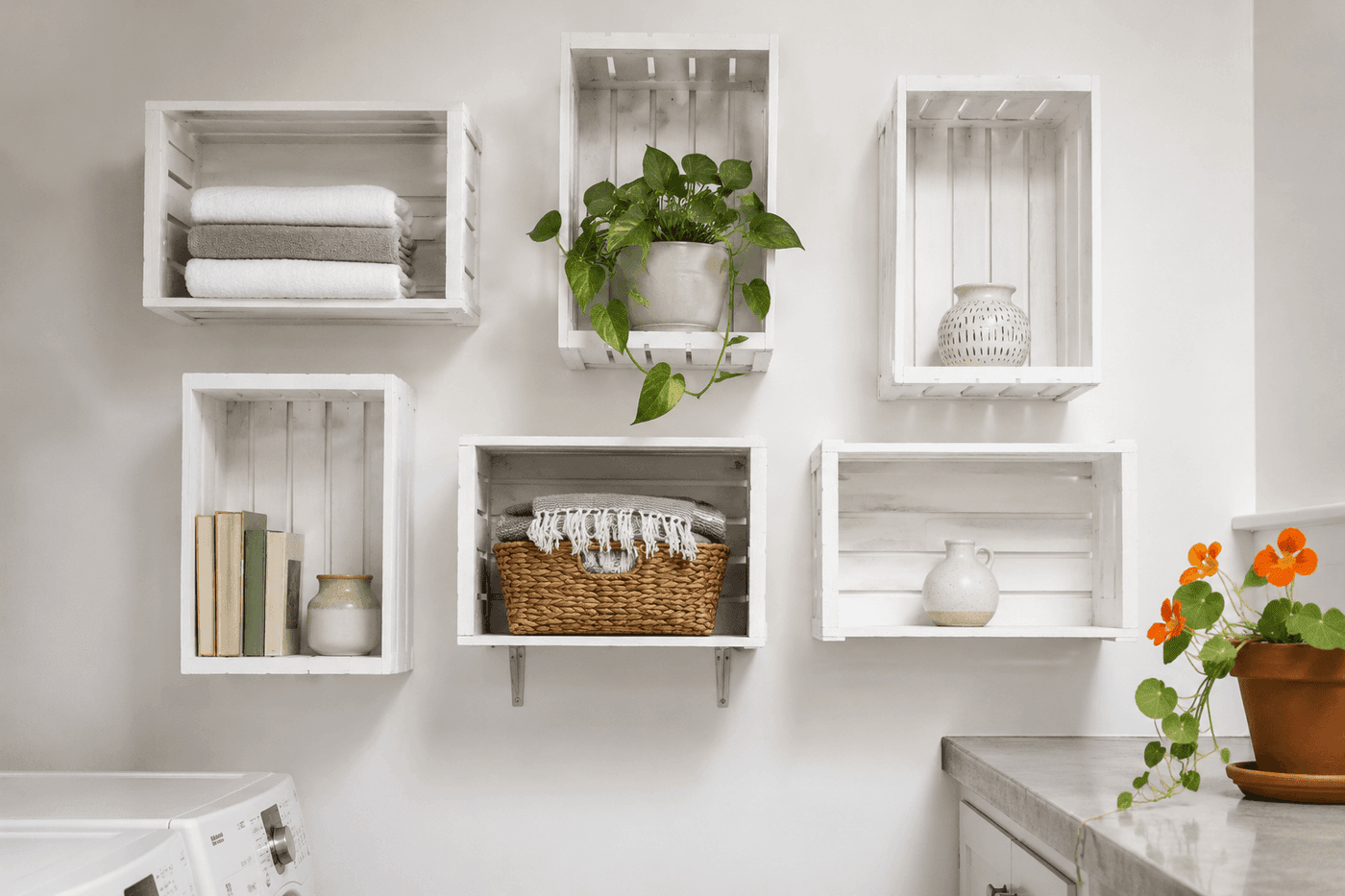 Six white chalk-painted wooden crates mounted on a laundry room wall in a staggered arrangement alternating horizontal and vertical orientations, filled with folded towels, small plants, and storage baskets