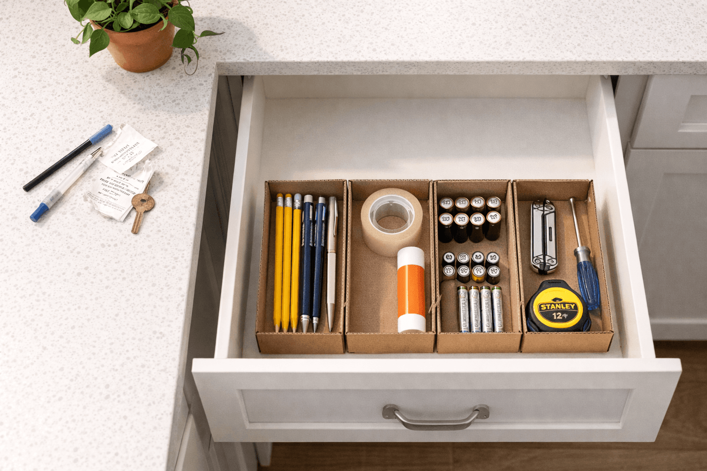 A neatly organized kitchen drawer divided into compartments with writing utensils, batteries, tape, and small tools grouped by category in repurposed small boxes and cardboard dividers